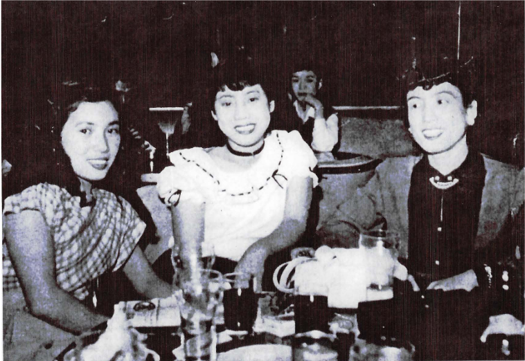 50s black-and-white image of 3 Japanese young women seated at a restaurant table smiling at the camera.